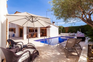 Zona junto a la piscina con mobiliario de mimbre, un área de estar cubierta y una piscina azul cristalina.