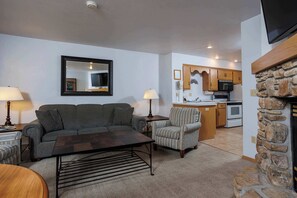 A living room with a sofa, armchair, coffee table, and lamps. A kitchen is visible in the background with wooden cabinets and appliances.