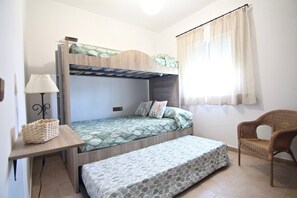 Un lit superposé avec un bureau et une chaise, une lampe et un fauteuil en osier, dans une pièce aux murs blancs et dotée d’une fenêtre avec des rideaux.