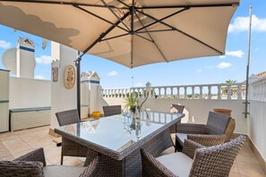 Un patio al aire libre con una mesa de vidrio, sillas de mimbre y un paraguas grande.