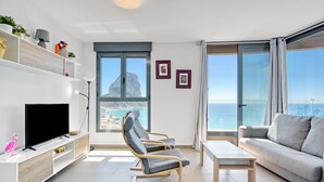 A modern living room with a TV, seating, a table, and a view of the sea.