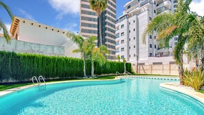 A swimming pool with a curved edge, surrounded by a low hedge, and adjacent to a multi-story building with balconies.
