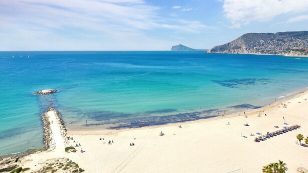A beach with clear blue water, a rocky breakwater, and a distant mountain.