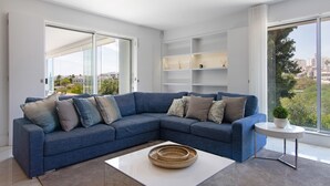 A modern living room with a blue sectional sofa, a white coffee table, and a view of the city through large windows. - Villa "Sweet Home Albufeira" with Private Pool, 2 Private Terraces & Wi-Fi (Albufeira)