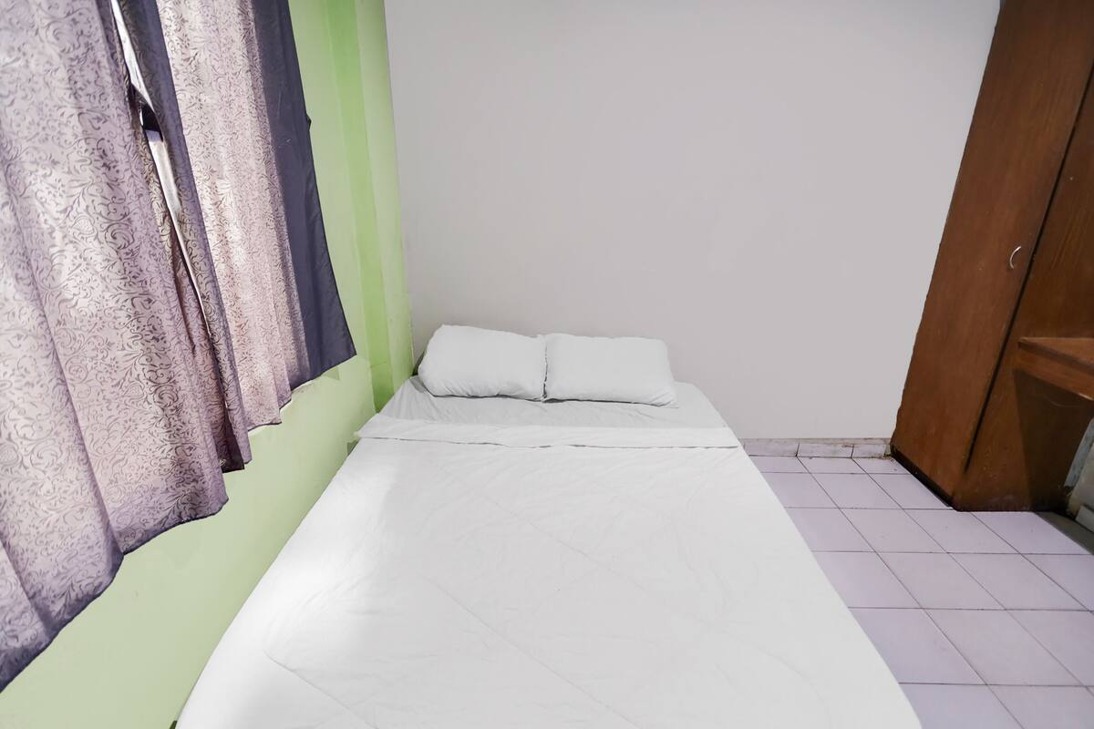 Habitación pequeña con cama individual, pared verde, ropa de cama blanca y puerta entreabierta.