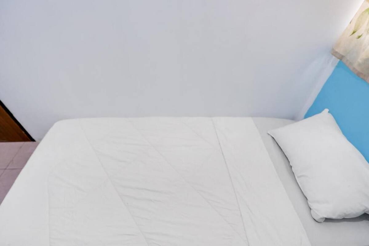 Una cama con colchón y almohada blancos, una pared azul y una ventana con cortina.