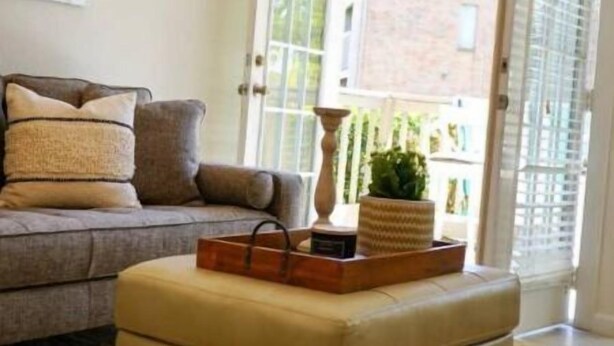 Una sala de estar con sofá, reposapiés, lámpara y ventana con vista a un edificio.