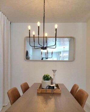 Un coin repas comprenant une table en bois, six chaises, un lustre, un miroir et un plateau avec une plante et une bougie.