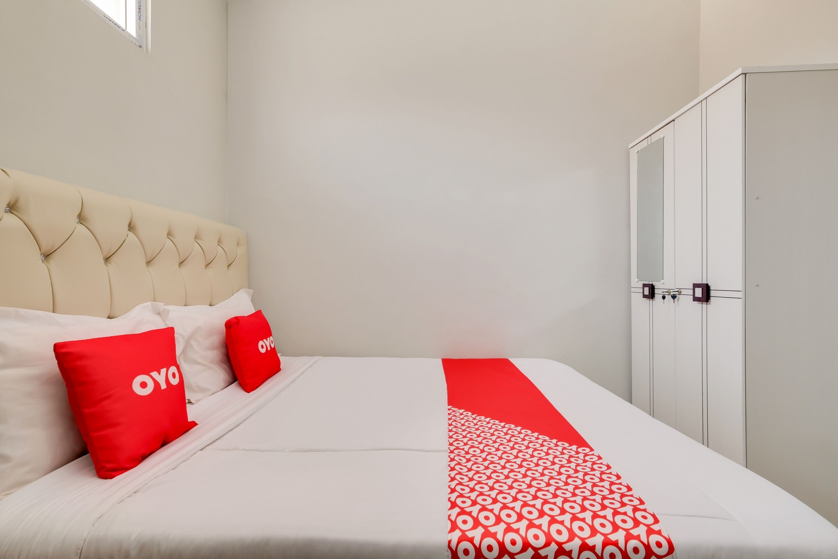 A hotel room with a bed, two red pillows, a white and red patterned bedspread, a beige upholstered headboard, and a wardrobe with a mirror.