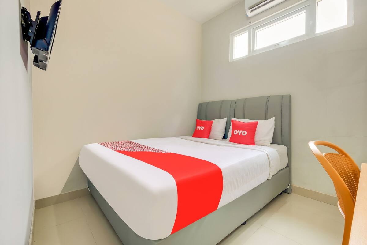 A neatly made bed with a red and white bedspread, two pillows with 'OYO' branding, a grey upholstered headboard, and a small table with a chair in the corner.