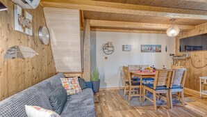 A cozy room with a wooden ceiling, a sofa with patterned cushions, a dining table with chairs, and a wall clock.