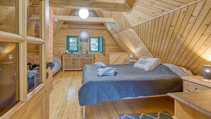 A cozy attic bedroom with a wooden sloped ceiling, a bed with a blue quilt, a nightstand, a dresser, and a window with a view of trees.