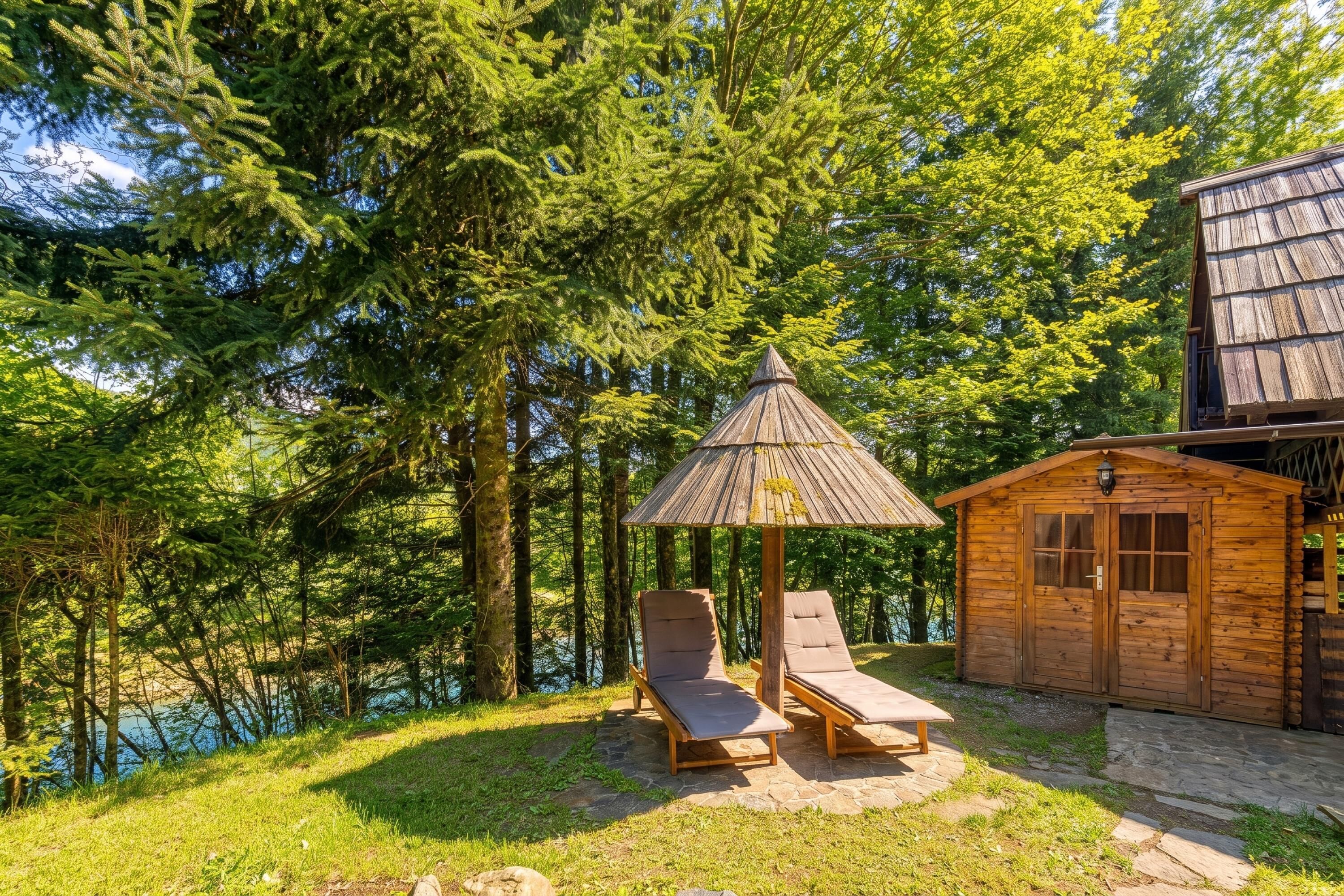 A wooden cabin with a thatched roof, two lounge chairs, and a small wooden structure with a window.