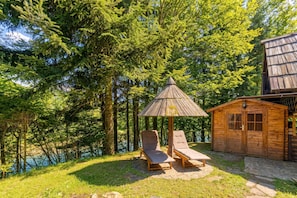 A wooden cabin with a thatched roof, two lounge chairs, and a small wooden structure with a window.