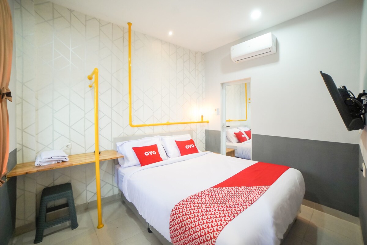 A hotel room with a bed, a desk, a mirror, and an air conditioning unit.