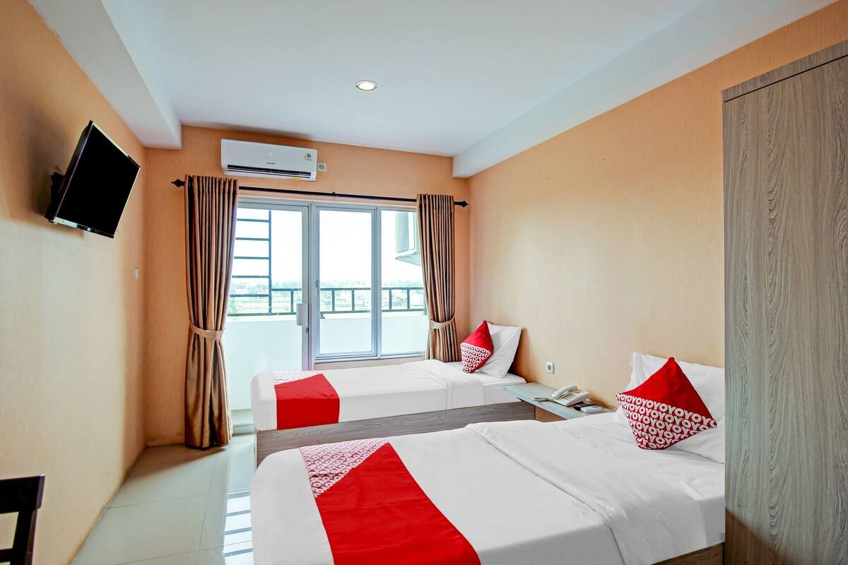 A hotel room with two beds, a TV mounted on the wall, an air conditioning unit, and a window with curtains.