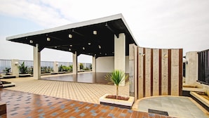 A covered outdoor area with a tiled floor, a palm tree planter, and a modern building with a sloped roof. - Hotel O Tridasa (Medan)