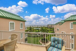 A balcony with wicker furniture, a view of a landscaped garden, and buildings with green roofs. - Oceanfront Condo Pool Hot tub Private Beach Access (Saint Simons Island)