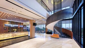 A modern hotel lobby with a marble reception desk, a curved staircase, and large windows offering views of the outdoors.
