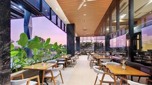 A spacious indoor dining area with wooden tables and white chairs, large windows offering views of the outside, and a modern ceiling design.