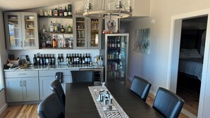 A dining area with a long table, chairs, a chandelier, and a bar area with shelves of bottles and glasses.