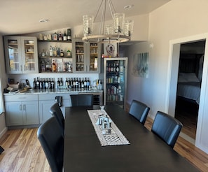 A dining area with a long table, chairs, a chandelier, and a bar area with shelves of bottles and glasses.