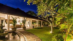 A well-lit outdoor area with a stone pathway leading to a covered seating area, surrounded by lush greenery and a small water feature. - Baliani Beauty (Seminyak)
