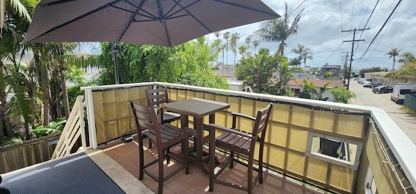 A balcony with a table and chairs, a large umbrella, and a view of palm trees and buildings.