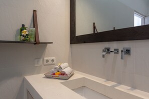 Ein Badezimmer mit einem weißen Waschbecken, einem Spiegel und einem Handtuchhalter mit Handtüchern. Zwei Steckdosen befinden sich an der Wand.