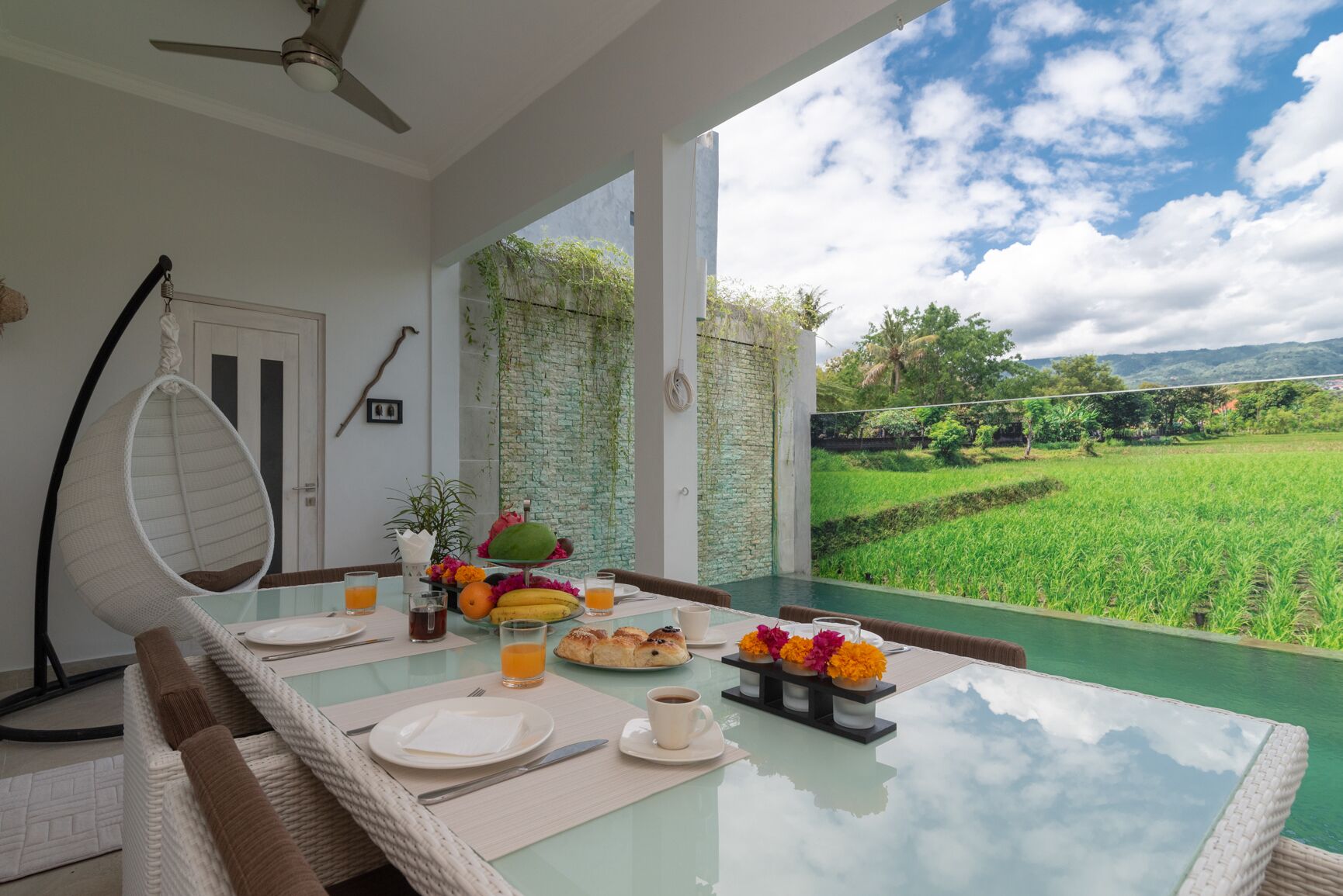 Una mesa de comedor dispuesta con platos, cubiertos y una cesta de frutas, con vistas a un campo verde exuberante.
