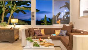 Een woonkamer met een bruine bank, decoratieve kussens, een salontafel met eten en drinken, een lamp en grote ramen met uitzicht op de lucht en planten.