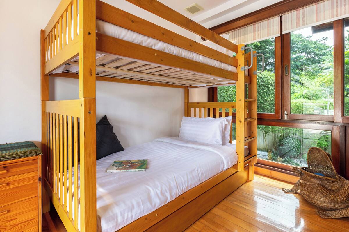 Uma cama beliche com estrutura de madeira, roupa de cama branca e janela com vista para o verde.