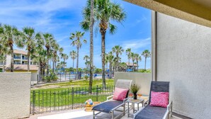 A balcony with two lounge chairs, a small table, and a view of a pool area with palm trees and a building in the background. - Stunning First Floor Condo, Direct Beach Access! Two Pools, One Heated Pool, $300 in Free Tickets! (St. Augustine Beach)