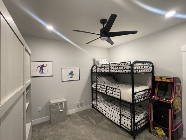 A bunk bed room with a ceiling fan, two framed pictures on the wall, a small trash bin, and a Pac-Man arcade machine.