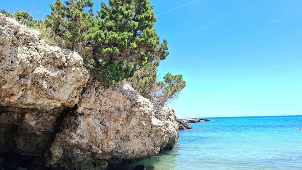 Una costa rocosa con aguas cristalinas, cielo despejado y un velero a lo lejos.