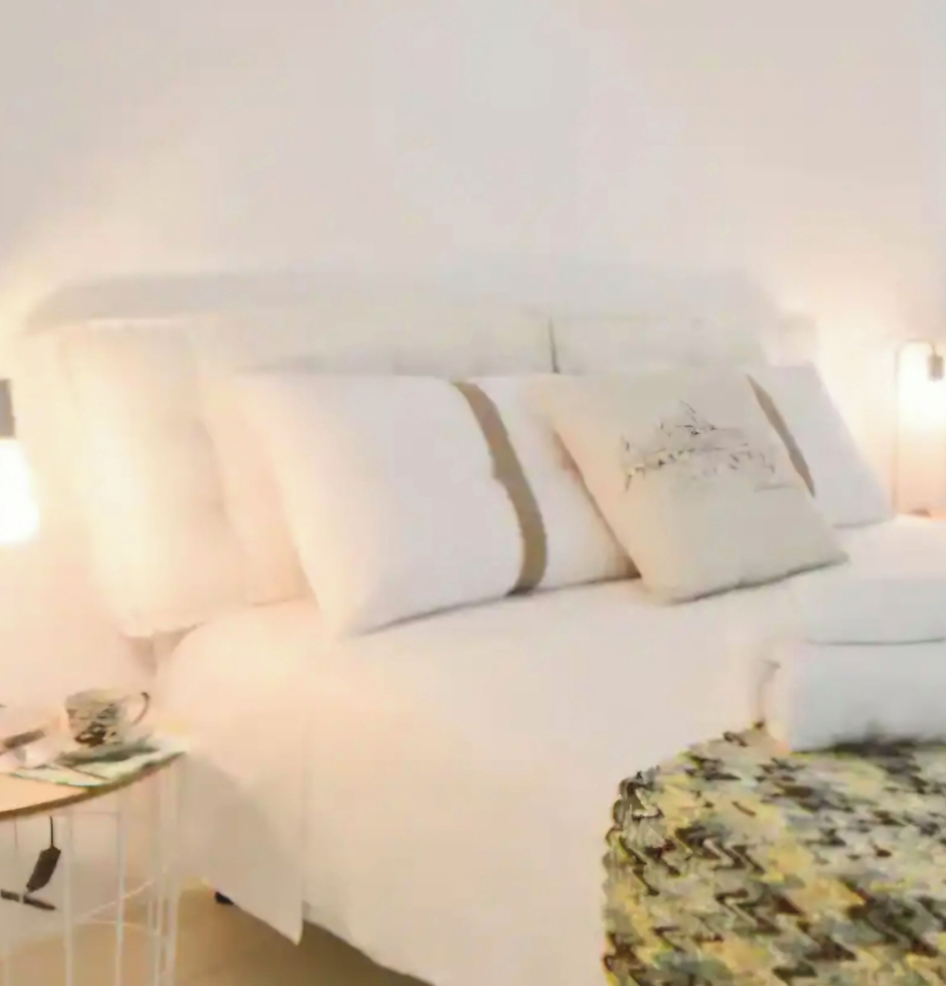A bed with white bedding and a patterned throw, a bedside table with a lamp, and a wall-mounted shelf.