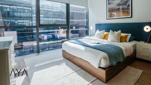 Ein modernes Schlafzimmer mit einem großen Bett, einem Nachttisch, einer Nachttischlampe und Blick auf eine Stadtlandschaft durch große Fenster.