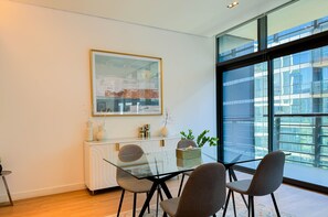 A modern dining area with a glass table, four chairs, a white console, and a large window with a city view. - Aya - Sophisticated 1BR Apartment in CityWalk with Views (Dubai)