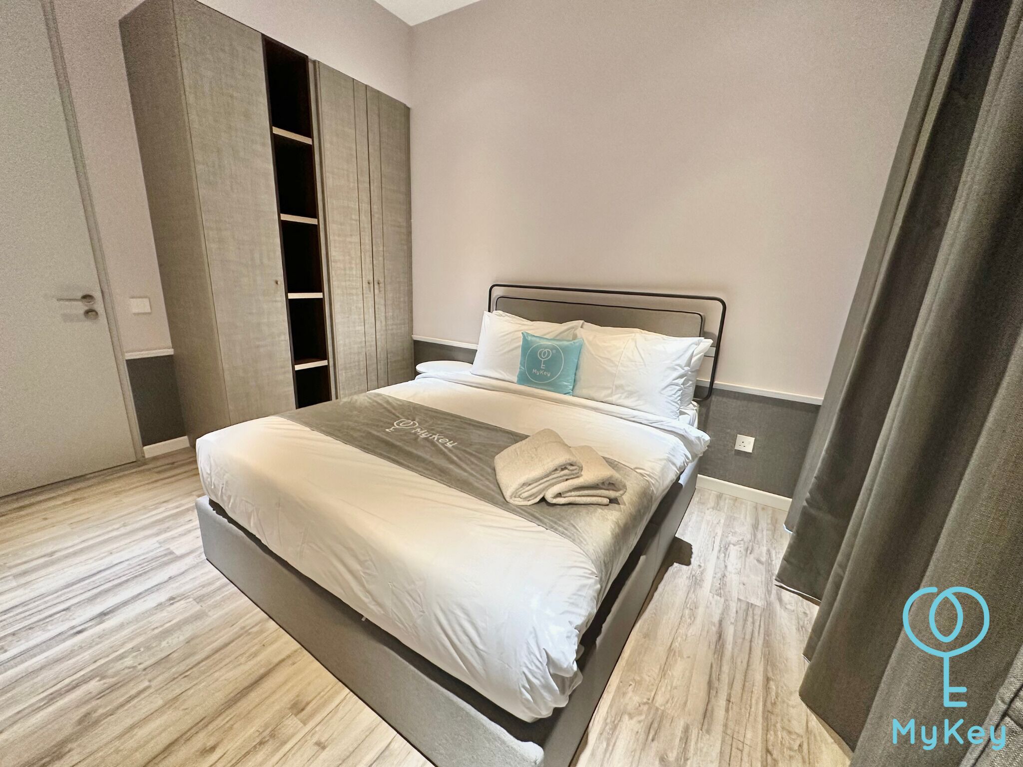 Een keurig opgemaakt bed met een grijze deken en een kussen met een logo, een garderobe met een spiegel en een grijze gordijn.
