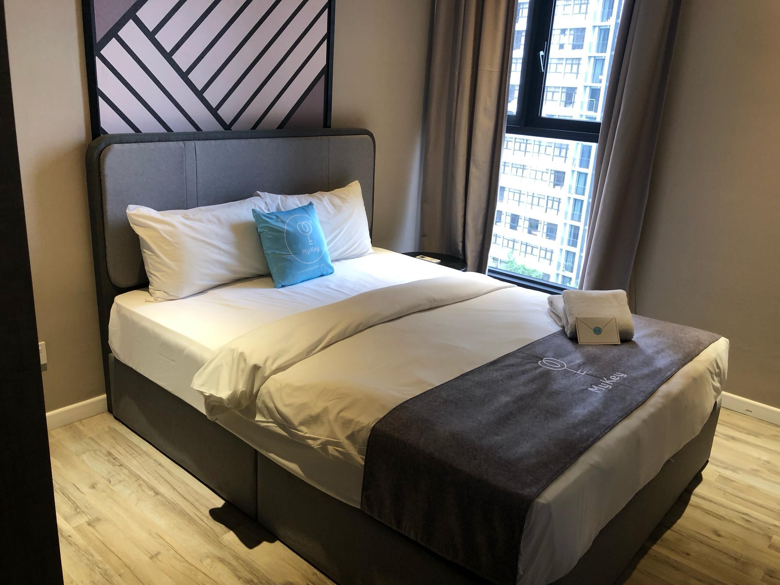 Una habitación de hotel moderna con una cama grande, un cabecero con diseño geométrico, vistas de la ciudad por la ventana y una pequeña mesa con una toalla.