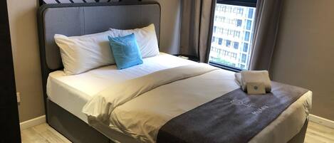 Una habitación de hotel moderna con una cama grande, un cabecero con diseño geométrico, vistas de la ciudad por la ventana y una pequeña mesa con una toalla.