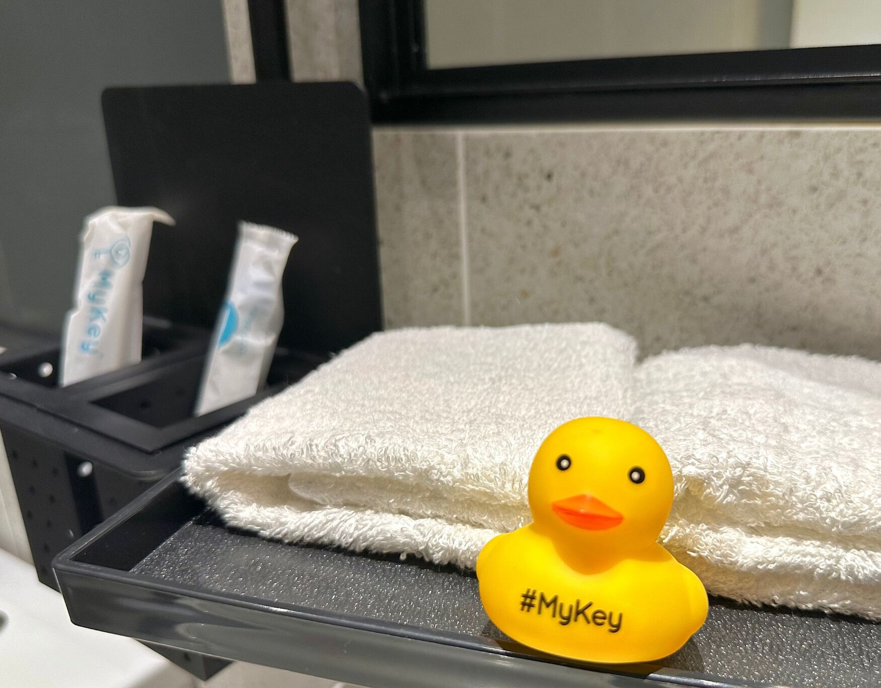 A bathroom with a white towel, a yellow rubber duck with #MyKey, and two tubes of cream.