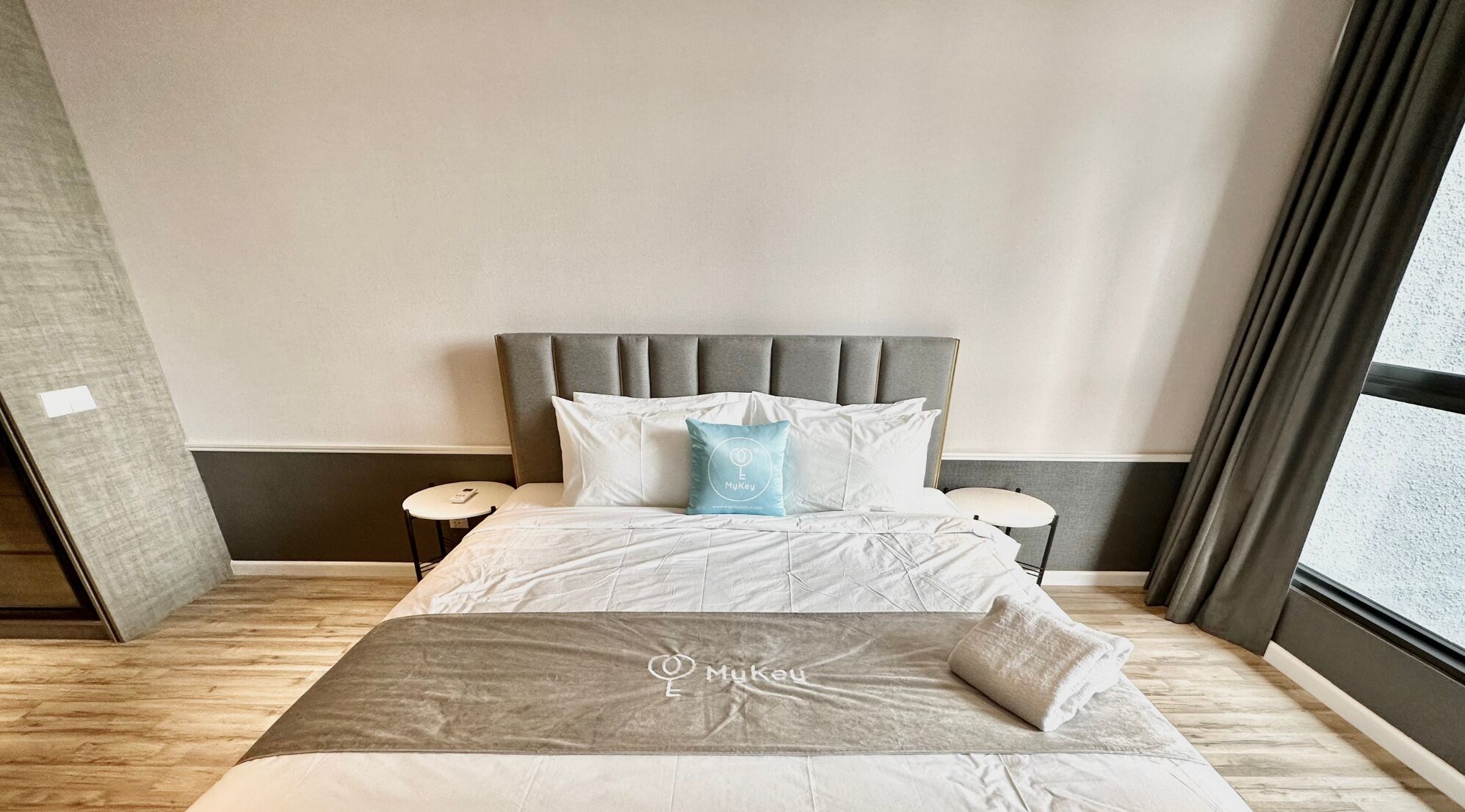 Une chambre d’hôtel moderne avec un grand lit, deux tables de chevet, une tête de lit rembourrée de couleur grise et une vue dégagée sur l’extérieur grâce à une grande fenêtre.