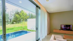 A modern living room with a large glass door leading to an outdoor swimming pool, a flat-screen TV on a wooden console, and a white sofa.