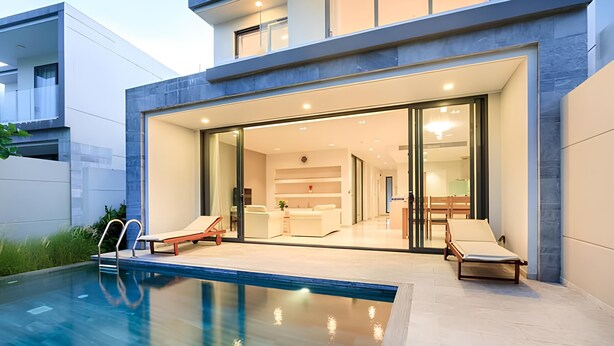 A modern house with a swimming pool, glass doors leading to a living area with a kitchen, and two lounge chairs by the pool.