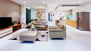 A modern living room with a sofa, ottoman, and coffee table, a kitchen area with a refrigerator, and a dining space with chairs and a table.