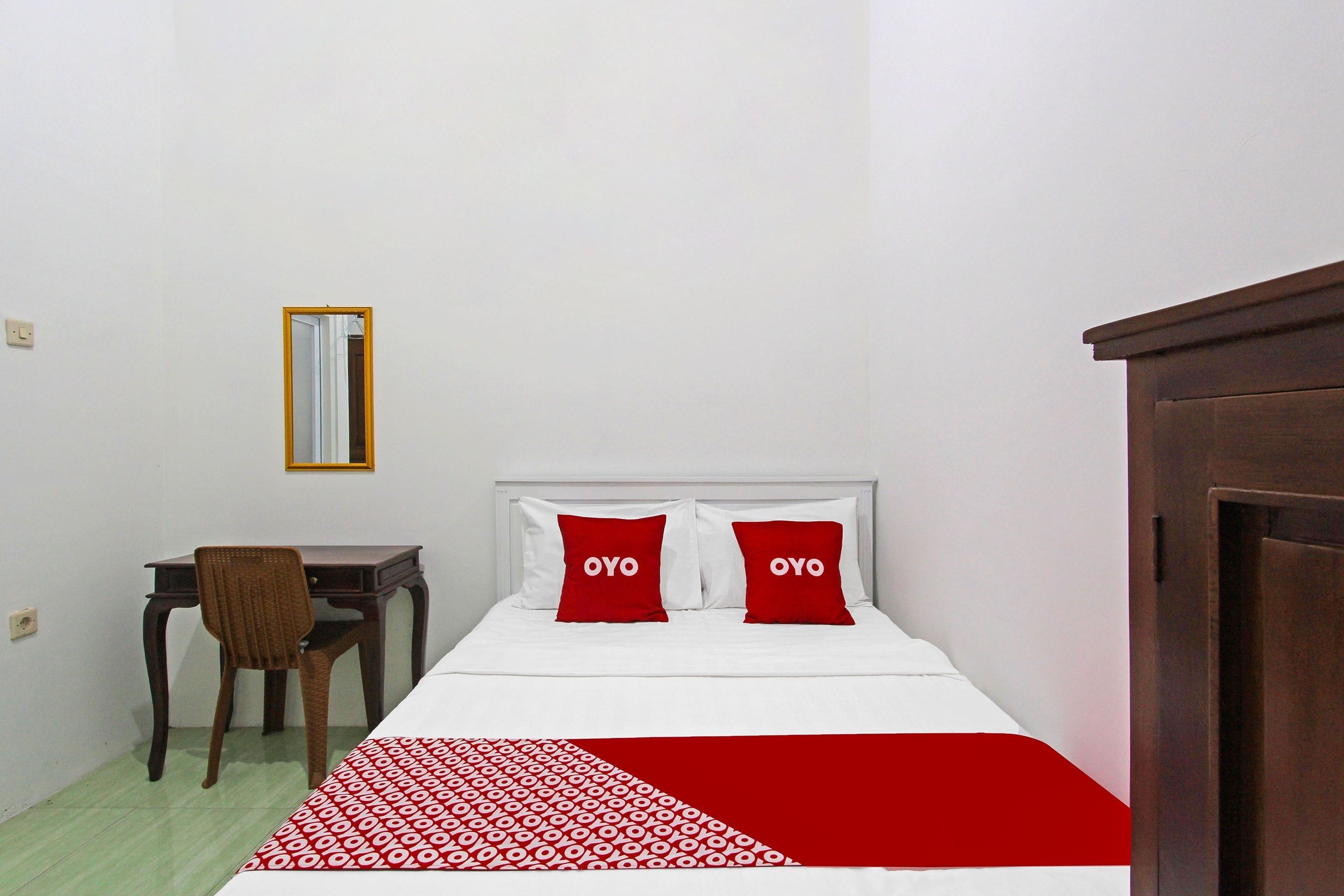 A bedroom with a bed, two red pillows with the word "OYO" on them, a wooden desk, a chair, and a mirror on the wall.