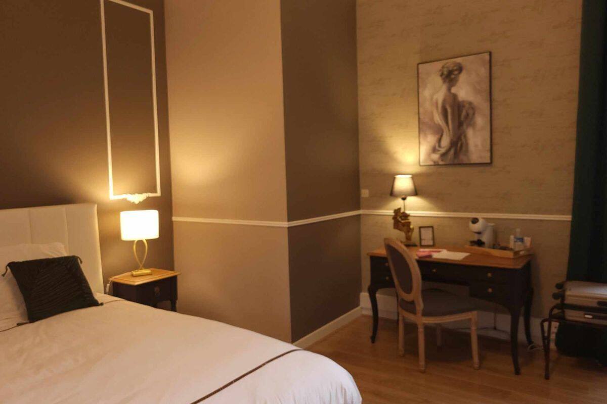 Quarto de hotel com cama, escrivaninha, cadeira, mesa de cabeceira e luminária na parede.