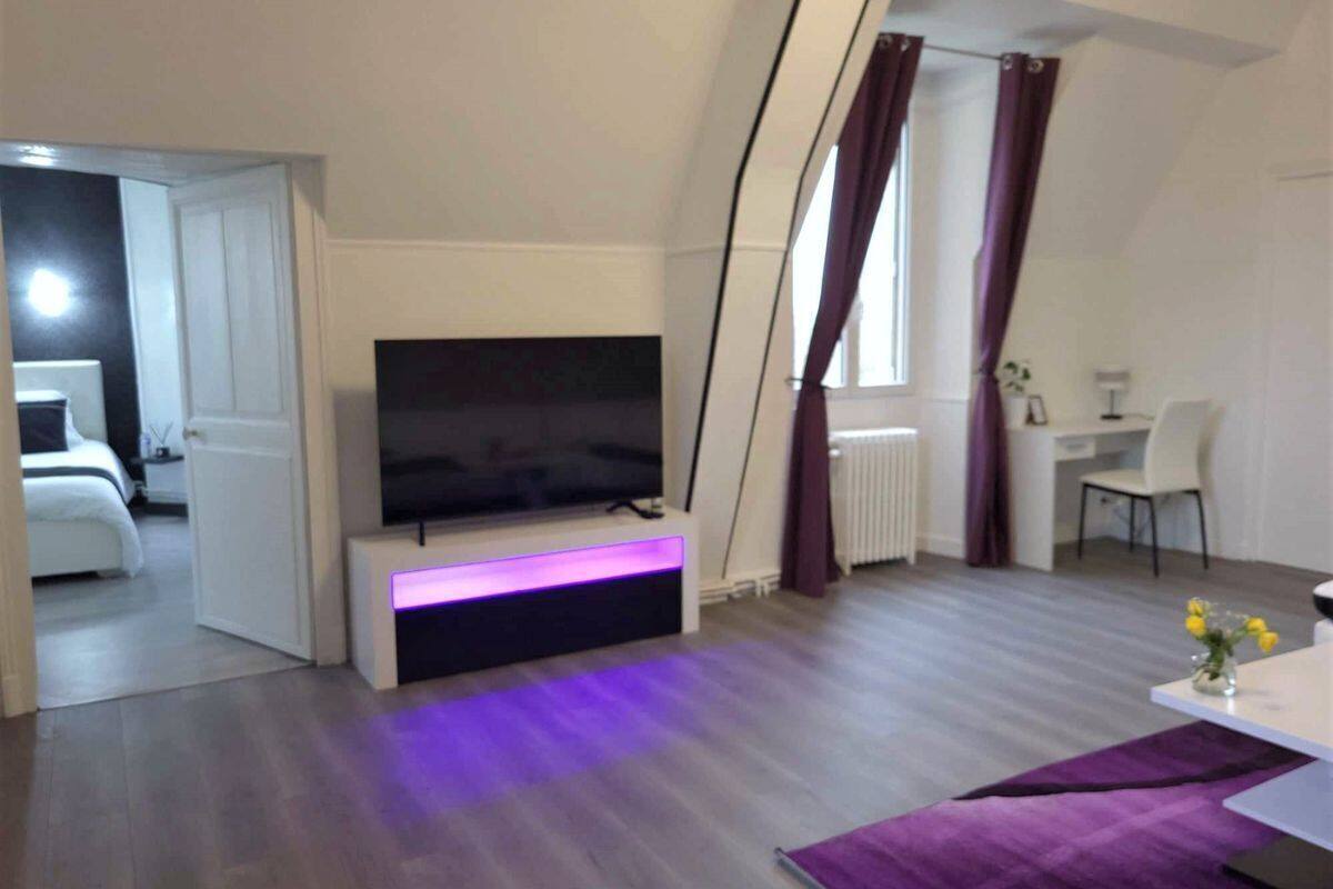 Uma sala de estar moderna com uma TV de tela plana em um móvel, uma área de jantar com mesa e cadeiras, e vista para um quarto por uma porta aberta.