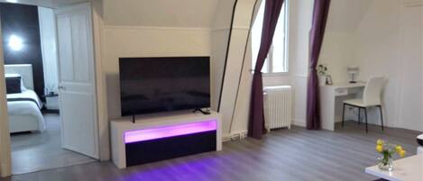 A modern living room with a flat-screen TV on a stand, a dining area with a table and chairs, and a view of a bedroom through an open door.